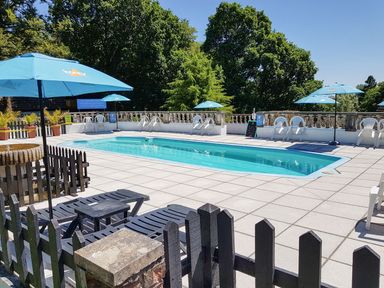 An outdoor swimming pool with blue umbrellas white chairs and black lounge chairs surrounded by trees at Valley Lodge 55 Callington near Gunnislake