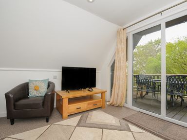 A sitting area with a black armchair with a floral cushion a wooden TV stand with a television and glass doors leading to a patio with outdoor furniture at Valley Lodge 55 in Callington near Gunnislake