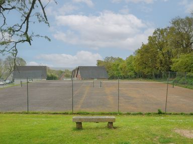 An outdoor tennis court with a stone bench on a grassy area surrounded by trees and houses at Valley Lodge 55 Callington near Gunnislake