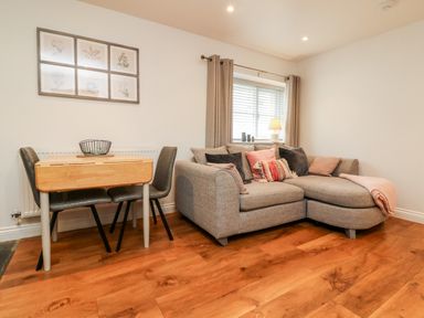 A living room with a grey corner sofa with cushions a small wooden dining table with chairs and a window with curtains at Cosy Cottage in Broadwoodwidger near Lifton