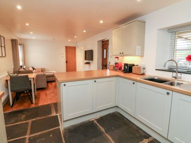 A kitchen with white cabinets a wooden countertop and a sink next to a living area with a sofa a table and chairs at Cosy Cottage Broadwoodwidger near Lifton