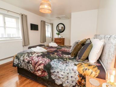 A bedroom with a bed covered in floral bedding pillows a wooden dresser a round wall mirror and two windows at Cosy Cottage in Broadwoodwidger near Lifton