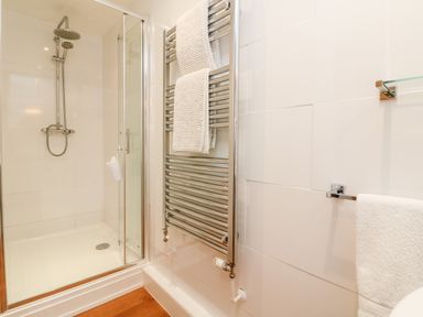 A shower area with glass door a chrome towel radiator with white towels and a white towel hanging on a rail in a bathroom at Cosy Cottage in Broadwoodwidger near Lifton