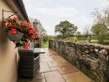A patio with a wicker chair and glass table with a grey umbrella and a hanging basket of red flowers with a stone wall and green fields beyond at Cosy Cottage in Broadwoodwidger near Lifton