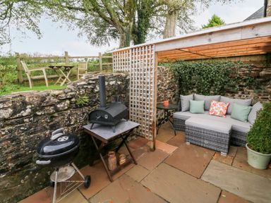 An outdoor patio area with a barbecue grill and pizza oven next to a stone wall and a seating area with cushions at Cosy Cottage in Broadwoodwidger near Lifton