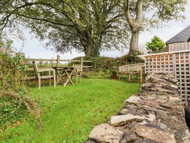 A garden area with wooden benches and table near large trees and a stone wall at Cosy Cottage in Broadwoodwidger near Lifton