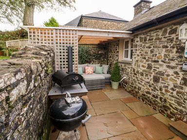 An outdoor patio area with stone walls a charcoal grill a pizza oven and a seating area with cushions at Cosy Cottage in Broadwoodwidger near Lifton