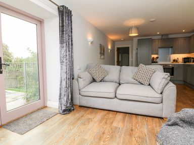 A living room with a grey sofa and patterned cushions near a glass door with grey curtains and an open kitchen in the background at The Old Cowshed Broadwoodwidger near Lifton