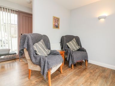 A room with two armchairs covered with gray throws and striped cushions a wooden floor a small wooden table a narrow bench near a door with vertical blinds and a wall light at The Old Cowshed in Broadwoodwidger near Lifton