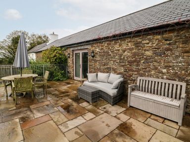 An outdoor patio with stone flooring a round wooden table with chairs a grey umbrella a curved cushioned sofa and a bench against a brick wall at The Old Cowshed in Broadwoodwidger near Lifton