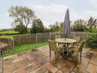 An outdoor patio with a round table and four chairs with a closed umbrella surrounded by a metal railing at The Old Cowshed in Broadwoodwidger near Lifton