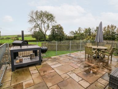 An outdoor patio with a grill table and a round table with chairs and an umbrella overlooking a green landscape at The Old Cowshed in Broadwoodwidger near Lifton