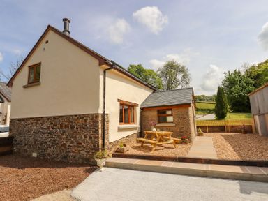 A cottage exterior with a wooden picnic table on a gravel patio and a garden in the background at Bunts Barn Northlew near Hatherleigh
