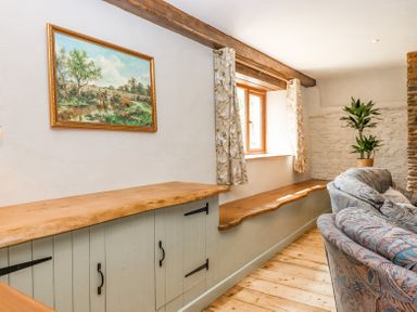 A room with wooden cabinets bench seating by a window patterned curtains a plant and a sectional sofa at Bunts Barn Northlew near Hatherleigh