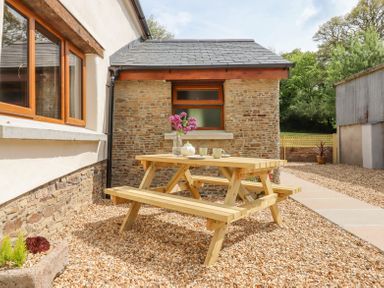 An outdoor area with a wooden picnic table set on gravel next to a stone and plaster building at Bunts Barn Northlew near Hatherleigh