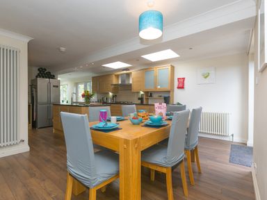 A dining area with a wooden table set with blue dishes and six chairs and a kitchen with wooden cabinets and stainless steel appliances at Spernenwyn in St Ives