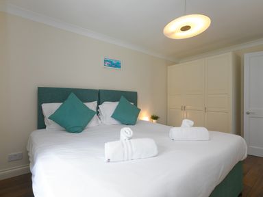A bedroom with a double bed white linens teal cushions rolled white towels wardrobe and a ceiling light at Spernenwyn in St Ives