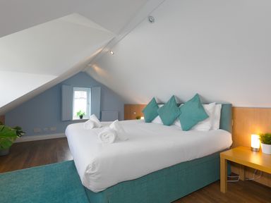 A bedroom with a large bed with teal cushions and rolled towels a wooden side table with a lamp and plant and a small window with shutters at Spernenwyn in St Ives