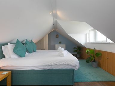A bedroom with a large bed with teal cushions and white bedding under a slanted ceiling at Spernenwyn in St Ives