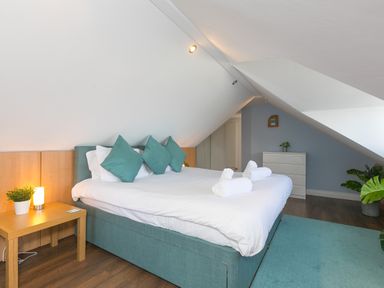 A bedroom with a double bed with teal pillows and white linens a wooden bedside table with a lamp and plant a white dresser and plants on a wooden floor at Spernenwyn in St Ives