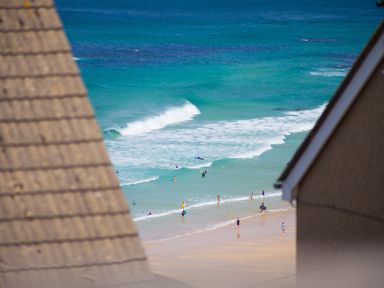 A beach with people swimming and walking near the ocean as waves come ashore framed by two rooftops at Spernenwyn in St Ives