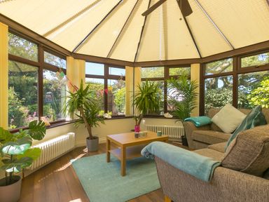 A conservatory with brown sofas a wooden coffee table plants and windows with vertical blinds at Spernenwyn in St Ives