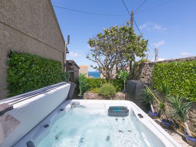 An outdoor hot tub surrounded by plants and a stone wall with a view of the sea at Spernenwyn in St Ives