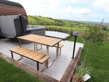 An outdoor patio with a wooden table and benches a hot tub a small tree and countryside views at Ceunant in Llanfihangel-y-Creuddyn near Aberystwyth