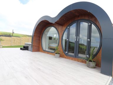 A curved wood and glass cabin with circular windows and doors on a tiled patio with potted plants at Ceunant in Llanfihangel-y-Creuddyn near Aberystwyth