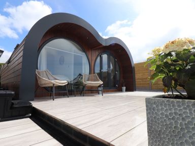 A wooden curved cabin with large round windows two chairs on a wooden deck and a potted plant at Ceunant in Llanfihangel-y-Creuddyn near Aberystwyth