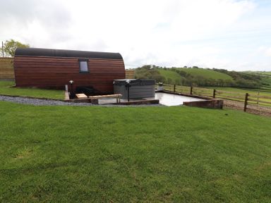 A wooden pod with a small window next to a hot tub on a patio surrounded by green grass and hills at Ceunant in Llanfihangel-y-Creuddyn near Aberystwyth
