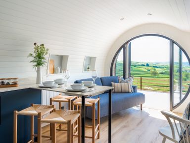 A kitchen and sitting area with stools bowls and glasses and a large round window showing green fields at Ceunant in Llanfihangel-y-Creuddyn near Aberystwyth