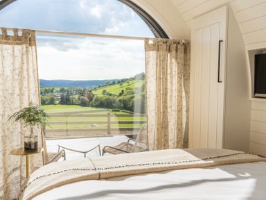 A bedroom with a bed a small table with a vase chairs and a large arched window overlooking green fields at Ceunant in Llanfihangel-y-Creuddyn near Aberystwyth