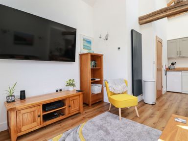 A living room with a yellow chair a wooden TV stand with plants a bookshelf and kitchen area at Harvenna Cottage in Crackington Haven