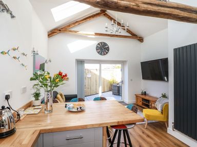 A kitchen and living area with wooden beams a dining table flowers on the table a yellow chair and a wall mounted TV at Harvenna Cottage in Crackington Haven
