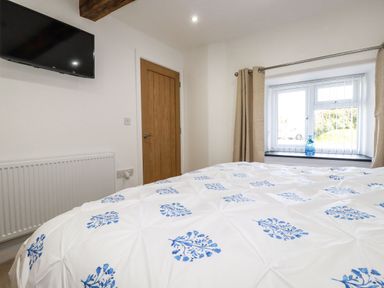 A bedroom with a bed covered in white and blue floral bedding a wooden door a wall-mounted television and a window with beige curtains at Harvenna Cottage in Crackington Haven