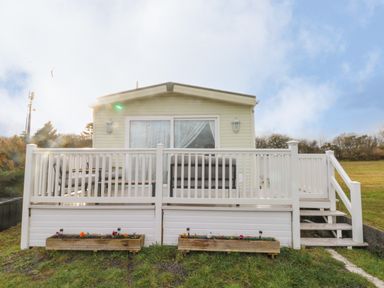A small beige house with a white wooden porch and railing in a grassy area at Park Lane in Llanteg near Kilgetty