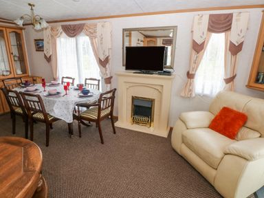 A dining area with a table set for six next to a fireplace with a television and an armchair with an orange cushion at Park Lane Llanteg near Kilgetty