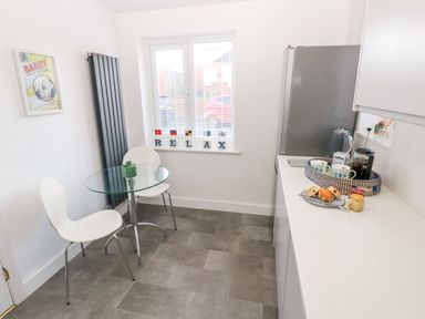 A small kitchen with a glass table and two white chairs next to a window with the word relax on the sill and a countertop with mugs and pastries at 12 Clos Yr Wylan in Barry