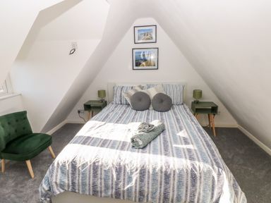 A bedroom with a bed striped bedding two round pillows two bedside tables with lamps a green chair and two framed pictures on the wall at 12 Clos Yr Wylan in Barry
