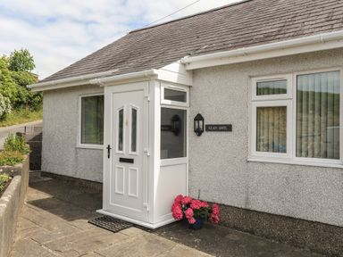 A front entrance with a white door and porch a potted plant with pink flowers on the ground near a window and a sign reading Glan Awel at Glan Awel in Nefyn