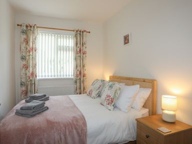 A bedroom with a double bed with floral pillows pink blanket towels on the bed window with floral curtains and a wooden nightstand with a lamp at Glan Awel in Nefyn
