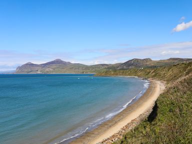 A coastal landscape with a sandy beach, blue sea, grassy cliffs, and hills in the background at Glan Awel in Nefyn