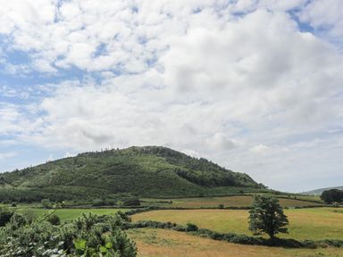 A grassy hill with trees and fields under a cloudy sky at Glan Awel in Nefyn