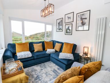 A living room with a blue corner sofa and mustard cushions by large windows at Pennant Cottage in Llithfaen