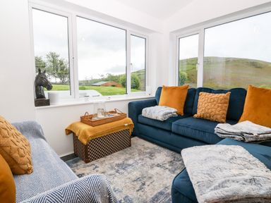 A sitting room with blue sofas and orange cushions overlooking a green landscape at Pennant Cottage in Llithfaen