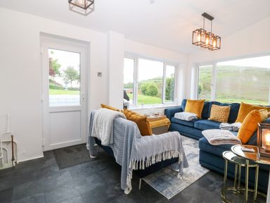 A living room with blue sofas and yellow cushions by large windows showing a green landscape at Pennant Cottage in Llithfaen