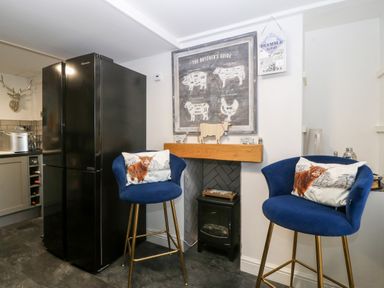 A kitchen corner with a black refrigerator two blue bar stools with animal pillows a small electric fireplace and a butcher's guide poster