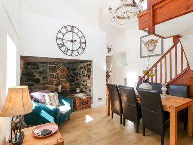 A dining area with a wooden table and black chairs next to stairs and a sitting area with a blue chair and a large clock on the wall at Pennant Cottage in Llithfaen