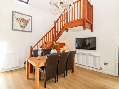 A dining area with a wooden table and six black chairs under a wooden staircase with a mounted television on the wall at Pennant Cottage in Llithfaen